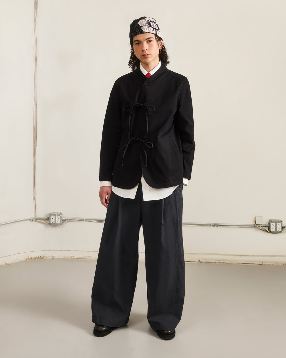 Indoors, a person wears the BLACK JEOGORI TIE JACKET by Sundae School over a white shirt, with wide dark pants, black shoes, and a patterned bandana. The plain background shows visible pipes and outlets along the wall.
