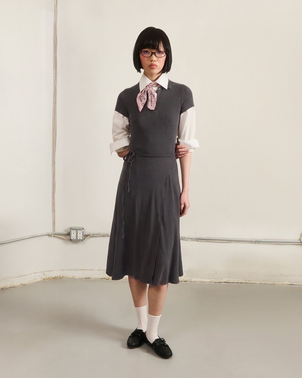 A person with short dark hair and glasses stands in a plain room with exposed wiring, wearing the Sundae School DARK GREY CINDY POLO DRESS layered over a white shirt, paired with a pink scarf, white socks, and black shoes.