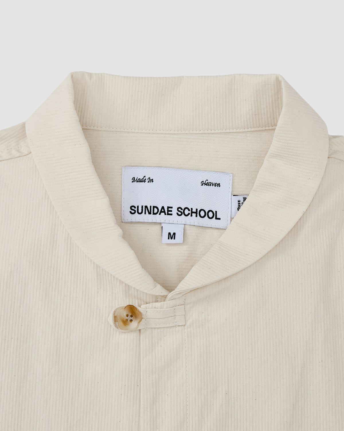 A close-up of the Sundae School IVORY CHINA COLLAR KNOT SHACKET, a cream-colored shirt with a mandarin collar and single button in corded cotton twill. The inner tag shows 