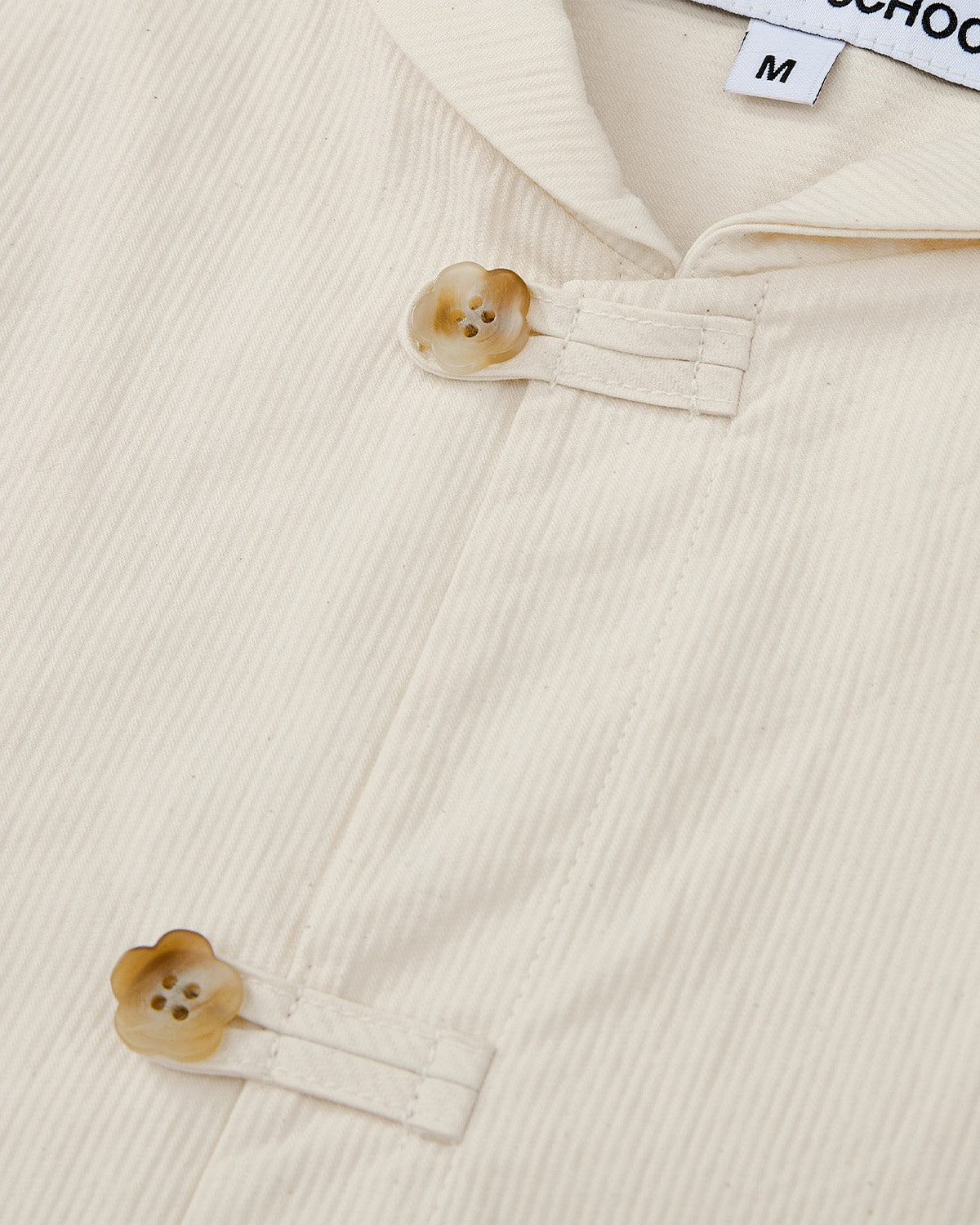 Close-up of Sundae School’s IVORY CHINA COLLAR KNOT SHACKET in corded cotton twill with vertical ribbing, two beige flower-shaped buttons, fabric loops at the collar, and a partial size M tag visible.