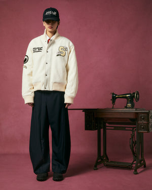 A person in the Sundae School IVORY VARSITY JACKET with vegan leather sleeves, loose dark pants, and a black cap stands beside a vintage Singer sewing machine against a plain magenta backdrop.