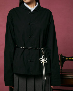 A person in a black buttoned jacket with a beaded belt featuring the Sundae School IVORY FLOWER KNOT KEYCHAIN stands before a pink background near a vintage sewing machine.