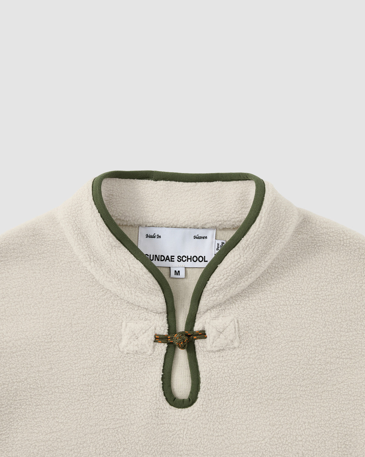 A close-up of the TAUPE TEARDROP KNOT FLEECE by Sundae School, featuring cream-colored fleece, olive green collar trim, and a loop-and-knot button closure at the neck; inside tag shows size M.