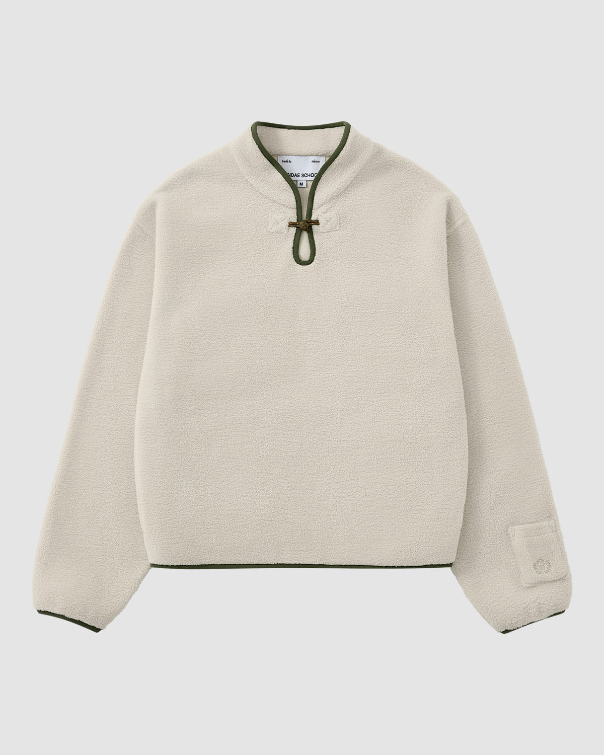 The TAUPE TEARDROP KNOT FLEECE by Sundae School is a cream-colored, long-sleeve pullover with a green-trimmed collar, front toggle closure, green edging on cuffs and hem, and a small patch detail on one sleeve.