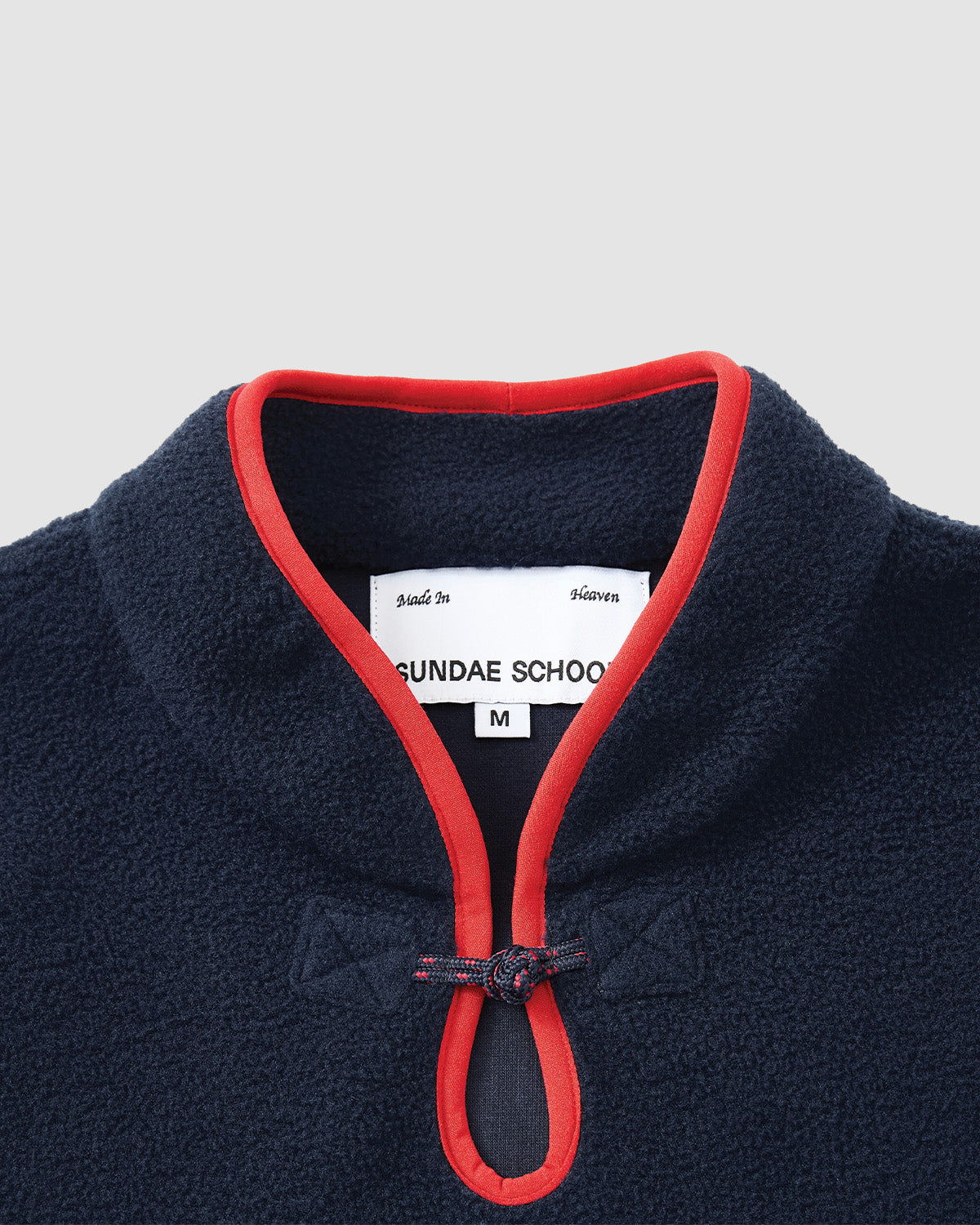 Close-up of the Sundae School NAVY TEARDROP KNOT FLEECE, a navy jacket with red-trimmed collar, knotted loop closure, and an inner white label marked SUNDAE SCHOOL M.