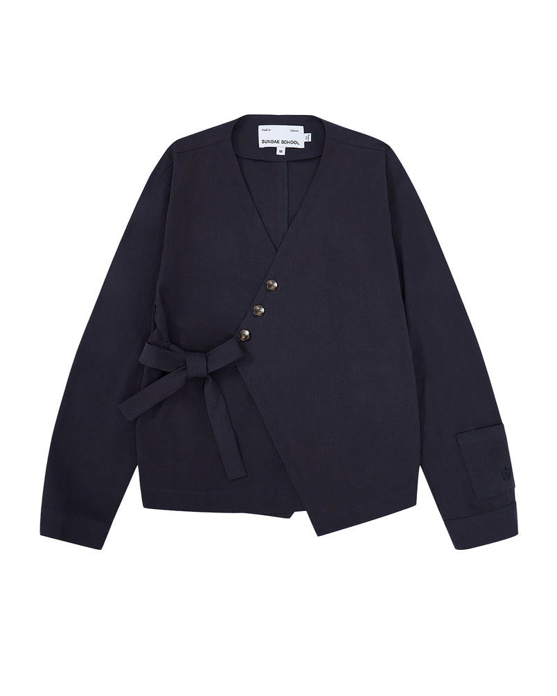 The NAVY HANBOK TIE BLAZER by Sundae School is crafted from 100% cotton in a navy blue hue, featuring a wrap-style silhouette with an asymmetrical front. It is detailed with a fabric belt for side tie closure, four buttons, and a single cuff accent on the sleeve.