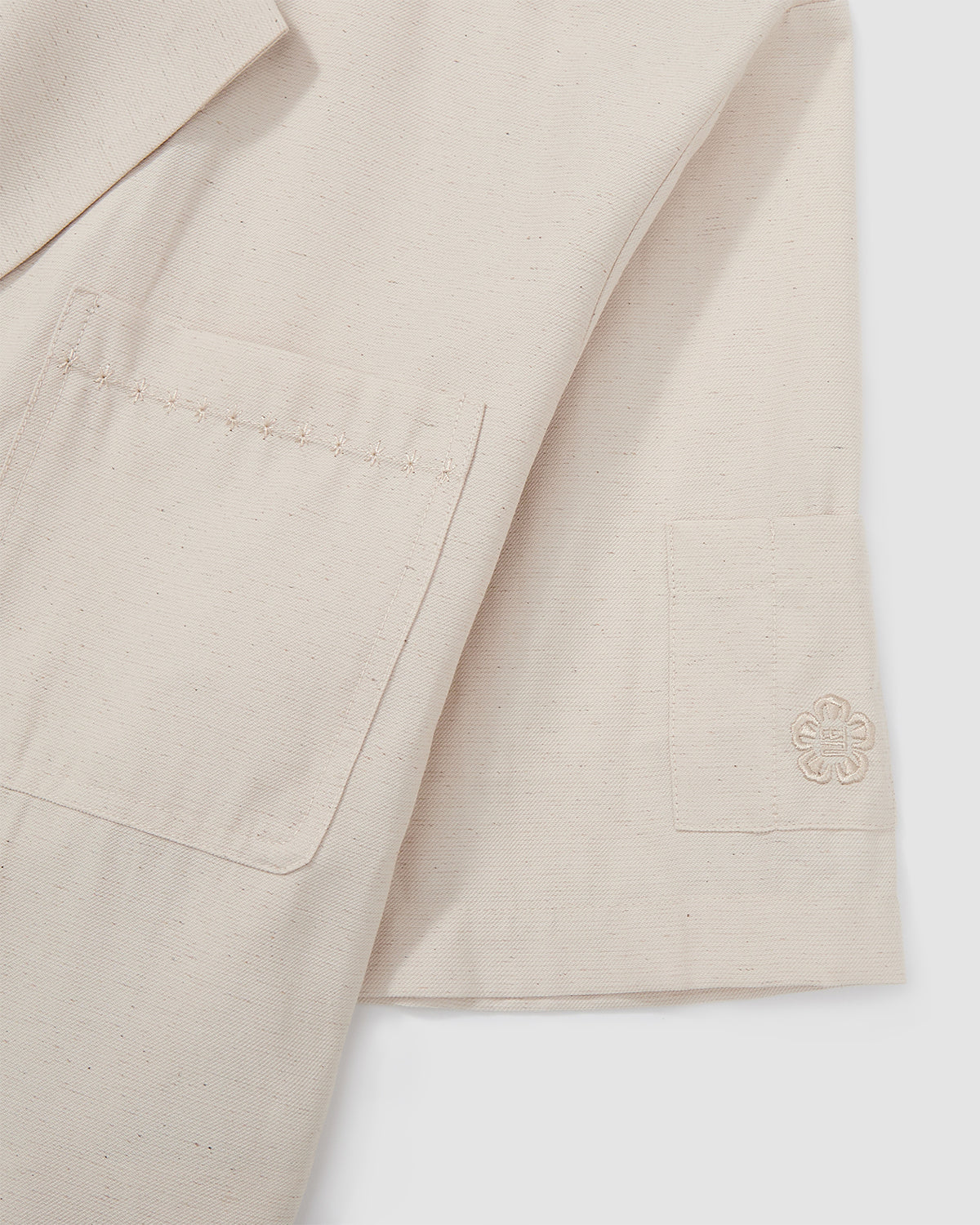 Close-up detail of cream hanbok shirt chest pocket with tonal topstitching and embroidered flower logo, showing construction details on textured linen blend fabric