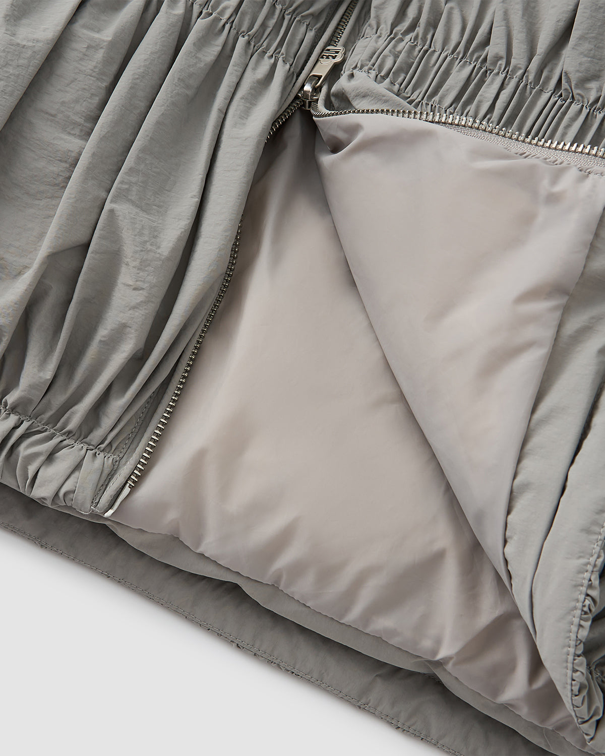 Close-up of Sundae School's SMOKE PLEATED DUVET PUFFER in light gray, showing the smooth interior lining, partially opened zipper, and gathered elastic seam details.