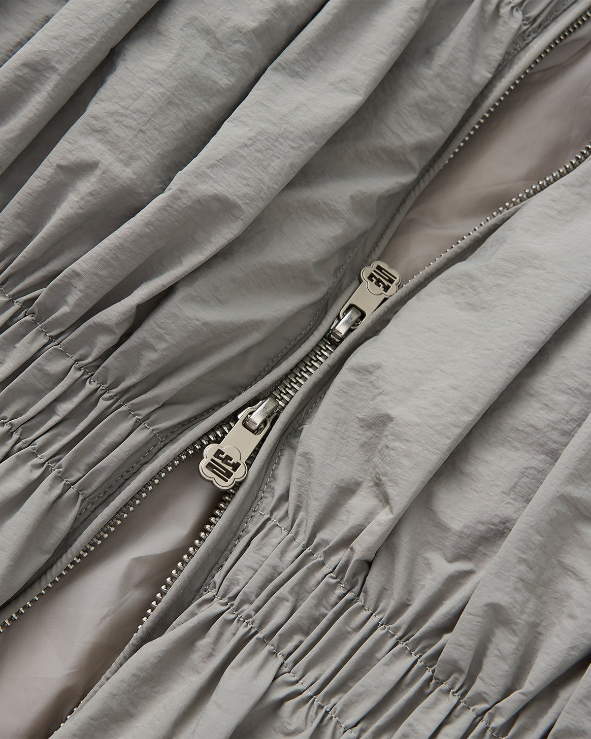 Close-up of a silver zipper partially unzipped on the light gray SMOKE PLEATED DUVET PUFFER by Sundae School, showcasing its ruched, cozy fabric reminiscent of a modern duvet coat.