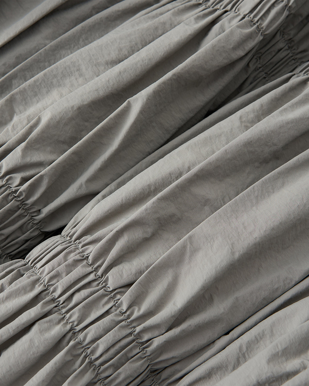 Close-up of Sundae School’s SMOKE PLEATED DUVET PUFFER shows light gray fabric with gathered, ruched sections and elasticized horizontal lines, creating soft, puffy folds for a cozy, stylish look.