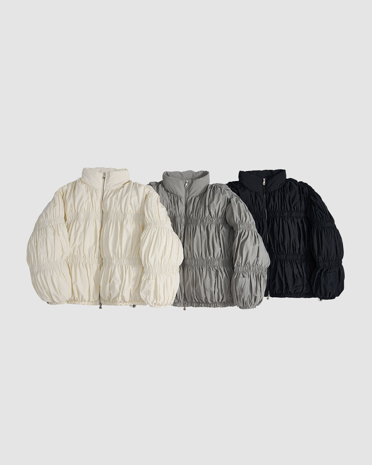 Three Sundae School SMOKE PLEATED DUVET PUFFER jackets with gathered, pleated fabric are displayed side by side in cream, gray, and black against a plain light gray background.