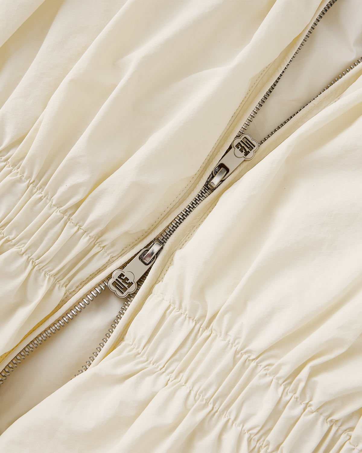 Close-up of a silver zipper on the lightweight, cream-colored IVORY PLEATED DUVET PUFFER by Sundae School. The slightly gathered fabric around the seams echoes the cozy, pleated design of this signature puffer.