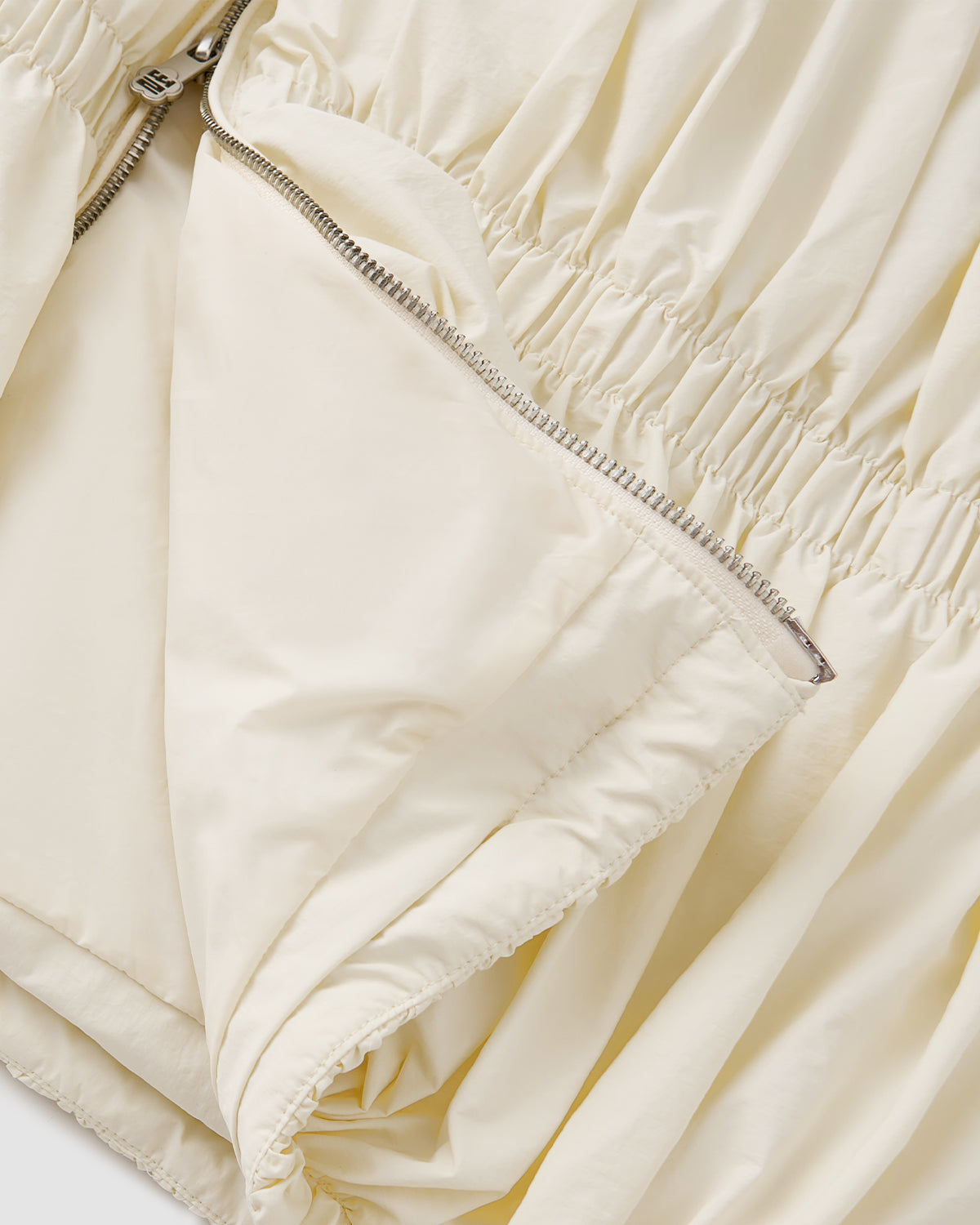 Close-up of Sundae School's IVORY PLEATED DUVET PUFFER, showcasing cream-colored pleated fabric, gathered elastic details, and a partially unzipped silver zipper that reveals the interior lining.