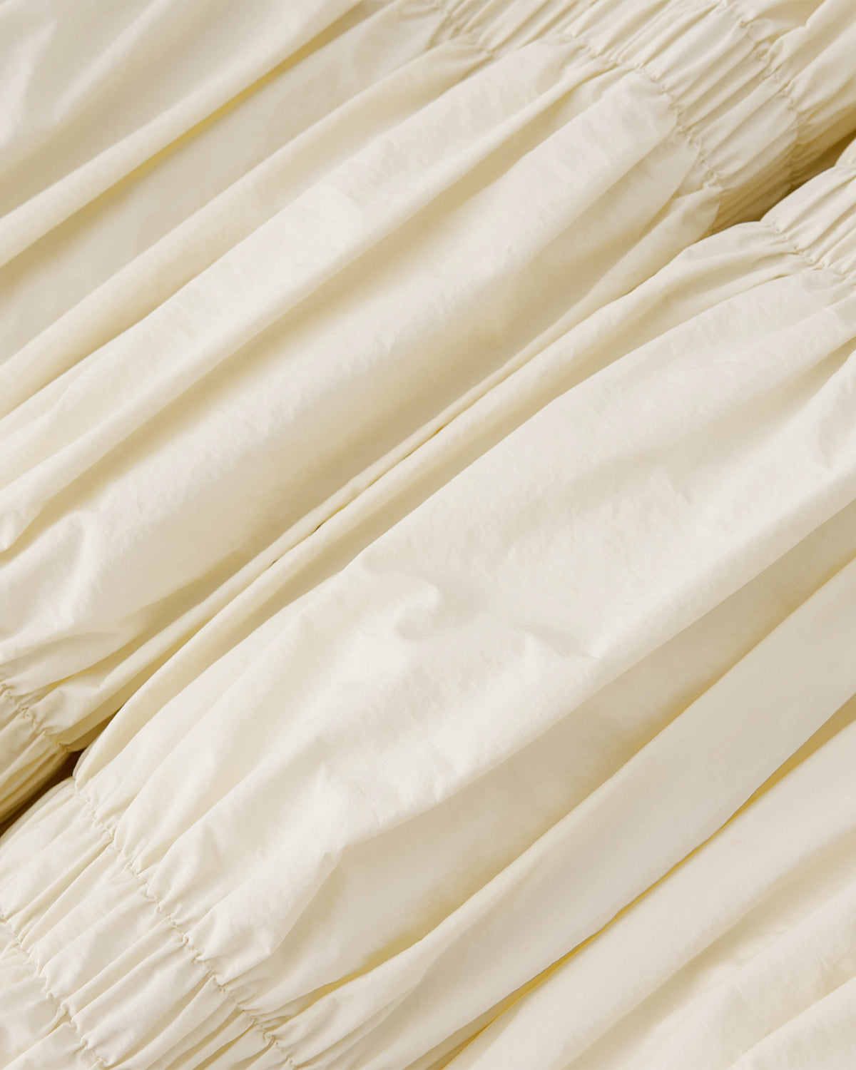 The IVORY PLEATED DUVET PUFFER by Sundae School features cream-colored fabric with elastic gathers, creating soft, ruched folds and parallel rows for a smooth, slightly shiny texture that evokes cozy, pleated duvet vibes.