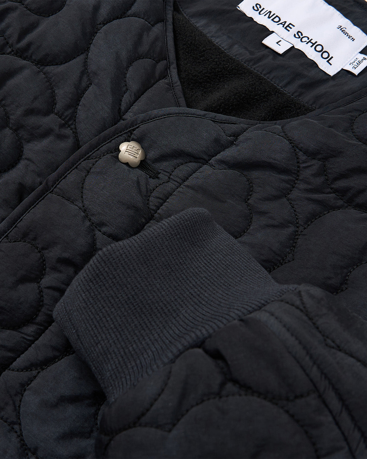 Close-up of the BLACK MOCHI QUILTED MILITARY LINER by Sundae School, featuring a ribbed cuff, metal button, visible quilted pattern, and size L label. This Korean military jacket is ideal for transitional weather.