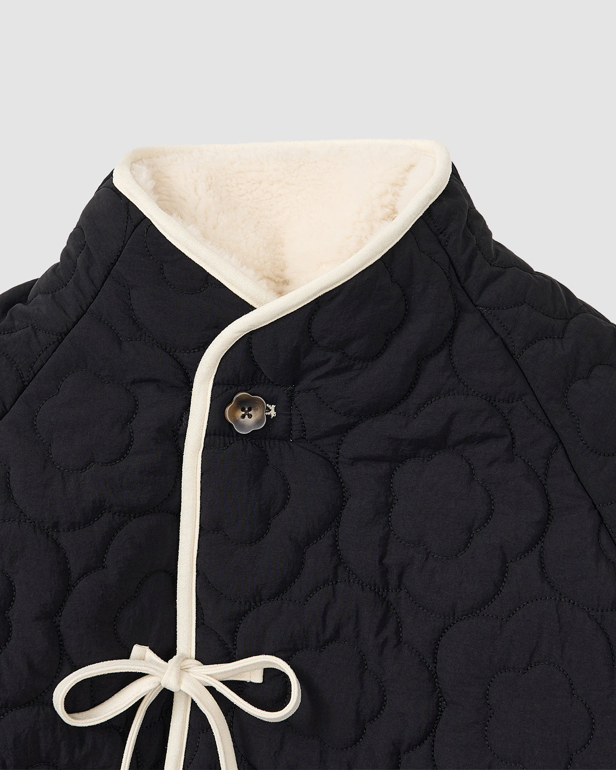 Close-up of Sundae School's BLACK IVORY REVERSIBLE FLOWER QUILTED JEOGORI FLEECE: a black Korean jacket with floral quilting, cream lining, off-white trim, single button, and front bow.