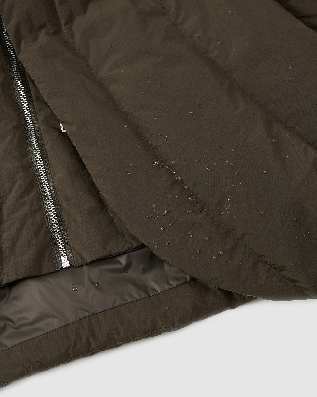 Close-up of the Sundae School KHAKI HEAVYWEIGHT MOCHI PUFFER, featuring water droplets beading on its water-resistant dark olive-green fabric. A silver zipper and part of the inner lining are also visible.
