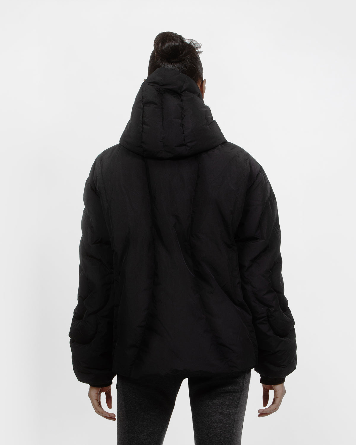 A person with dark hair in a bun models the Sundae School BLACK HEAVYWEIGHT MOCHI PUFFER, showcasing the back view and hood, paired with dark gray pants against a plain white background.