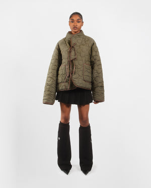 A person stands against a white background wearing Sundae School’s [PRE-ORDER] REVERSIBLE FLOWER QUILTED JEOGORI FLEECE in olive green, paired with a black pleated mini skirt, black knee-high leg warmers, and pointed-toe black shoes.