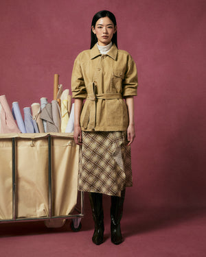 A woman poses before a mauve backdrop in the TAN ASYMMETRIC WORK JACKET by Sundae School, paired with a plaid skirt and black boots. Next to her, a beige cart overflows with pastel fabric rolls.