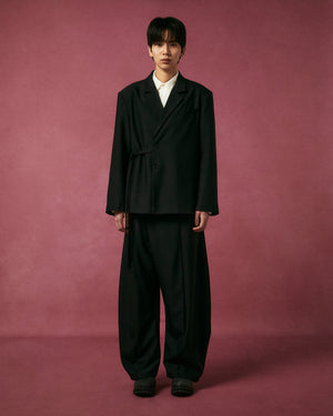 A person stands against a pink textured backdrop, wearing Sundae School’s BLACK PLEATED HANBOK SLACKS with wide legs, paired with an oversized black suit and white shirt, looking directly at the camera with a neutral expression.