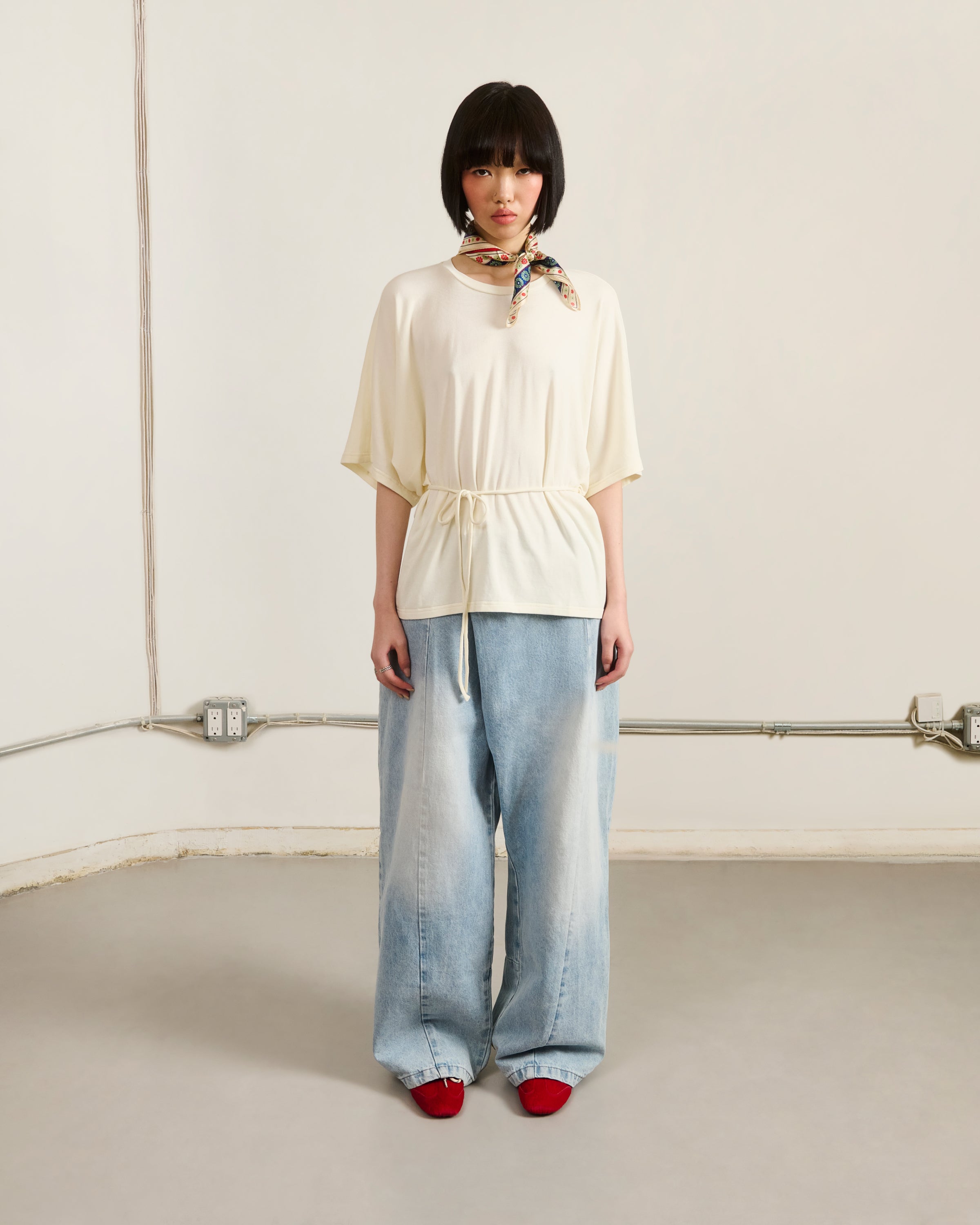 A person with short dark hair wears Sundae School SKY BLUE HANBOK DENIM PANTS, a cream belted top, red shoes, and a patterned scarf around their neck, standing against a plain wall with exposed electrical outlets and wires.