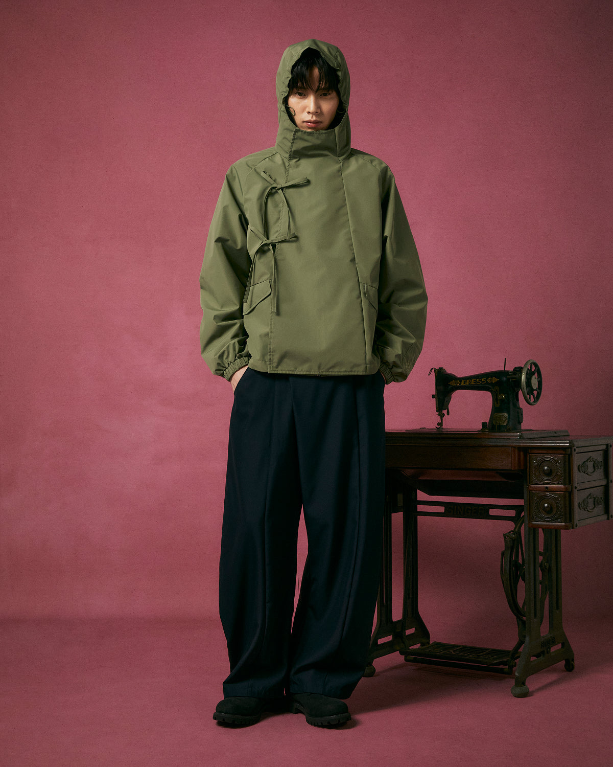 A person wears the MATCHA JEOGORI TIE SHELL JACKET by Sundae School with wide black pants, standing before a vintage sewing machine against a pink background.