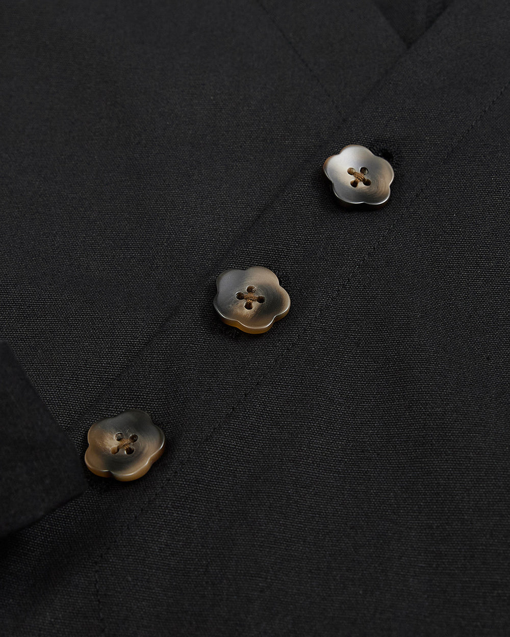 A close-up of the BLACK HANBOK TIE BLAZER by Sundae School showcases its black fabric adorned with three marbled brown and cream flower-shaped buttons, each featuring four holes and sewn in a vertical row—an elegant detail inspired by traditional hanbok jeogori silhouettes.