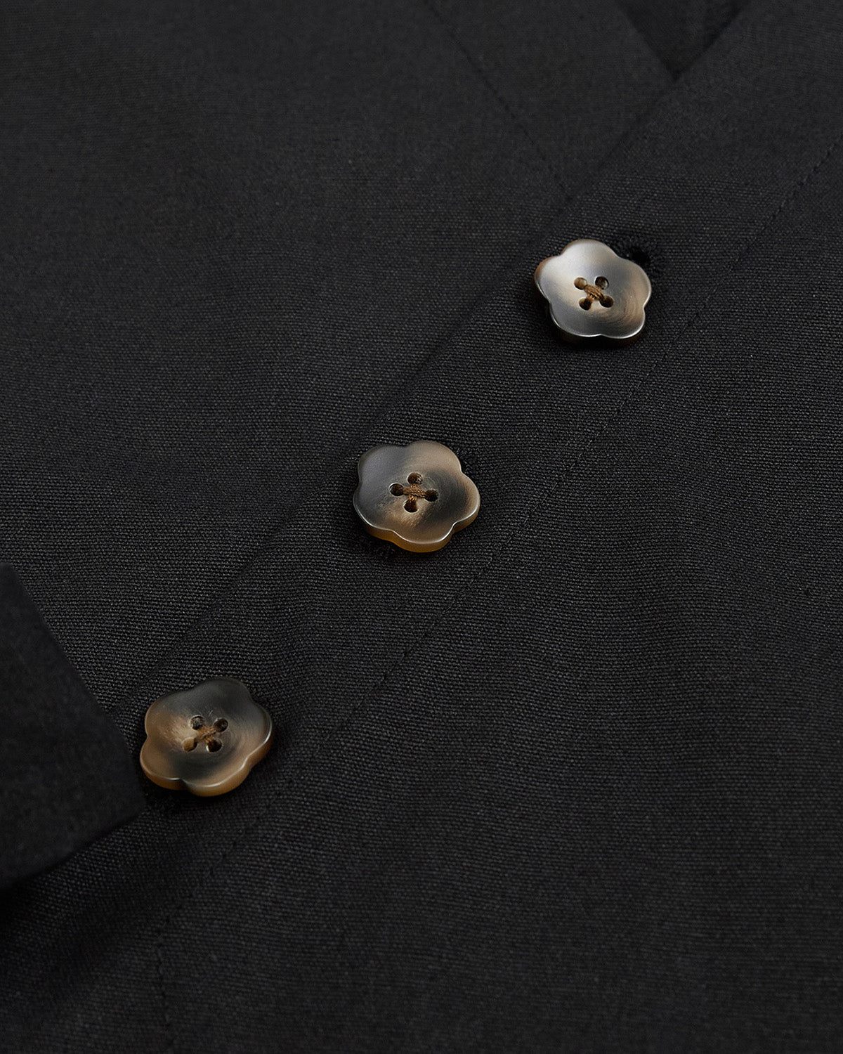 A close-up of the BLACK HANBOK TIE BLAZER by Sundae School showcases its black fabric adorned with three marbled brown and cream flower-shaped buttons, each featuring four holes and sewn in a vertical row—an elegant detail inspired by traditional hanbok jeogori silhouettes.