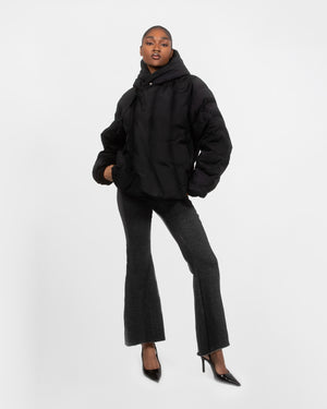 A person poses confidently in the BLACK HEAVYWEIGHT MOCHI PUFFER by Sundae School, paired with dark flared pants and black high heels against a plain white background.