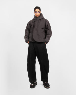 A person stands against a plain white background wearing the Sundae School CHARCOAL HEAVYWEIGHT MOCHI PUFFER, loose black pants, black platform shoes, and purple-tinted sunglasses with hands in their pockets.