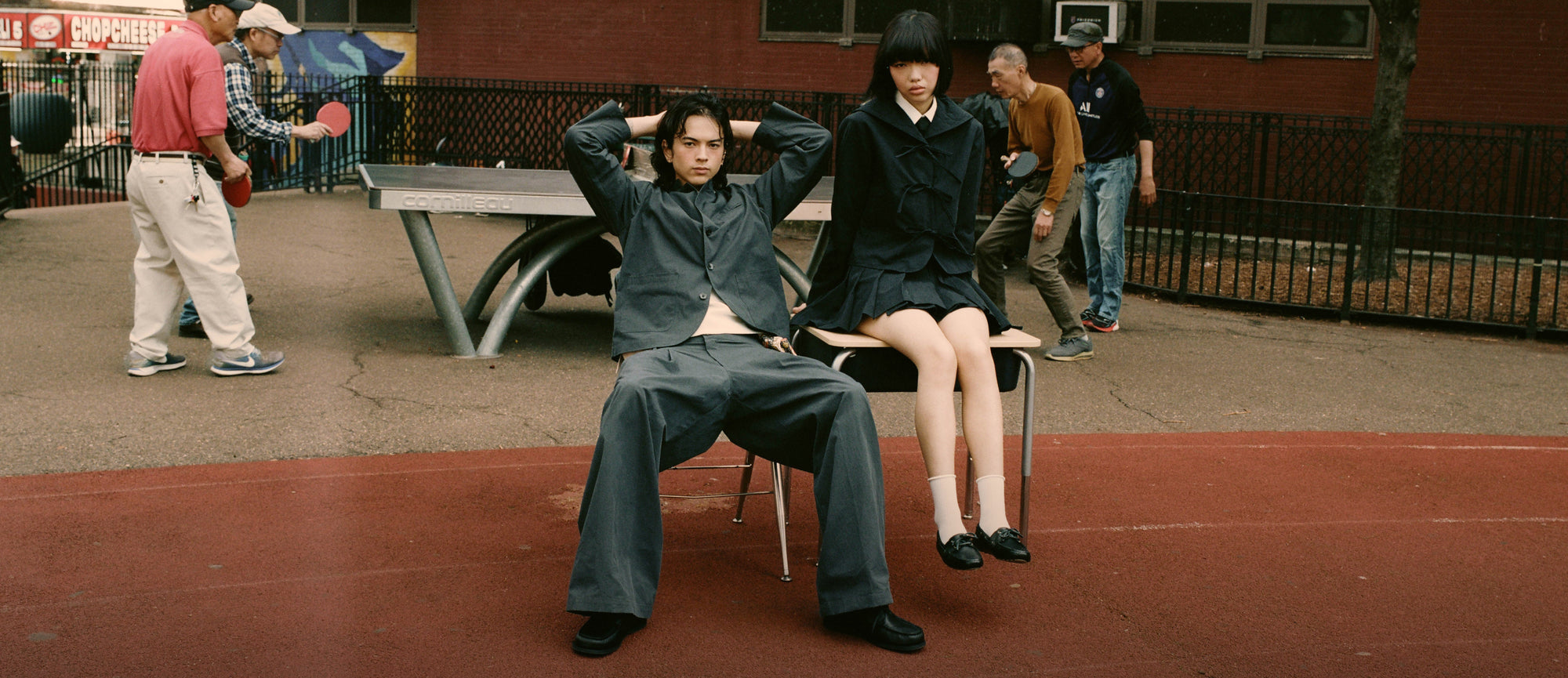 Two people sitting on chairs in a schoolyard with other students in the background wearing Sundae School Stand Collar Uniform Jacket and Pleated Uniform Slacks, Sailor Collar Uniform Jacket, and Uniform Apron Skirt 