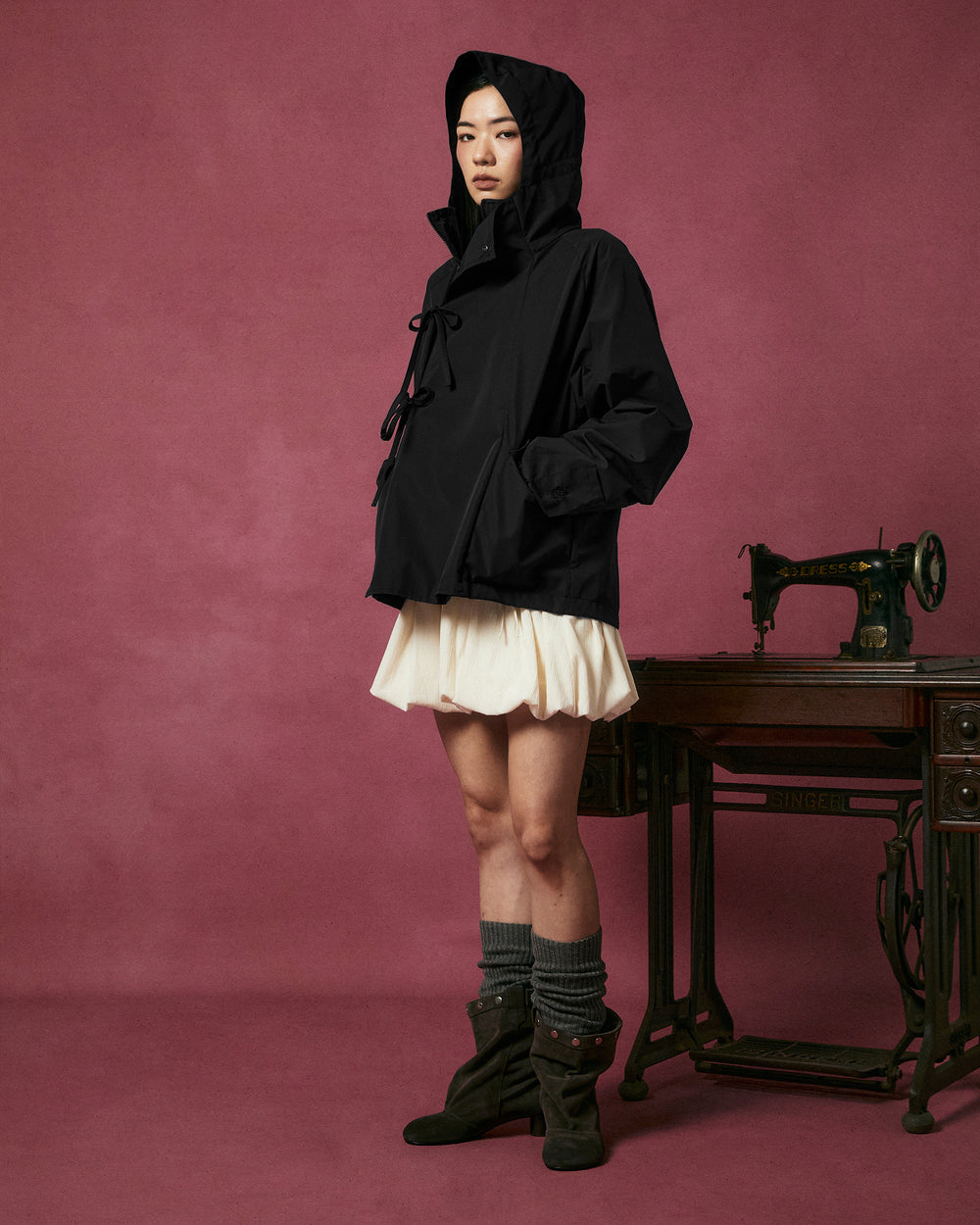 A person wearing the Sundae School BLACK JEOGORI TIE SHELL JACKET—a black, water-resistant hooded outerwear piece inspired by the Korean jeogori—paired with a white skirt, gray leg warmers, and brown boots stands next to an antique sewing machine against a pink background.