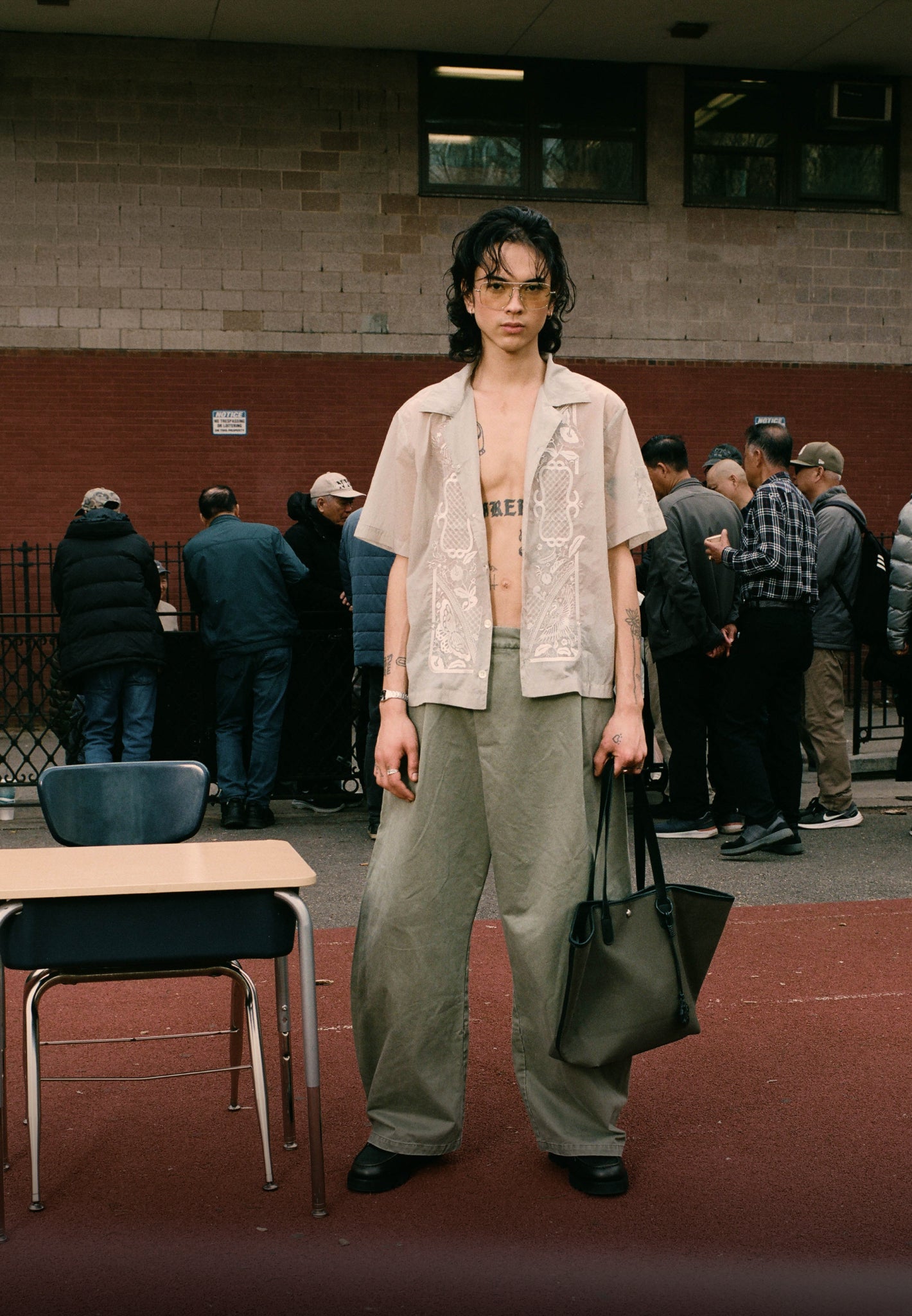 A vertical portrait of a model with long dark hair and glasses wearing an open, sheer embroidered cream shirt and wide-leg olive trousers. They are holding a large olive green tote bag, standing in an outdoor setting with a school desk in the foreground and a group of people in the background.