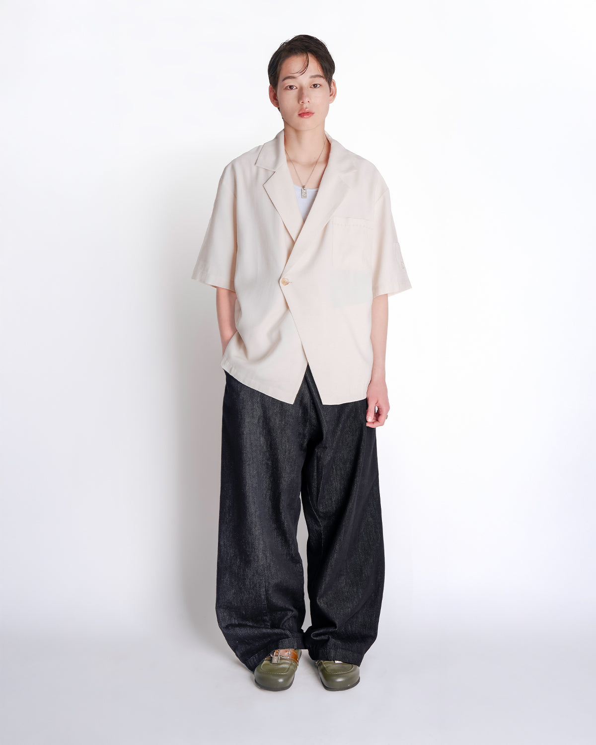 Cream hanbok-inspired short sleeve shirt modeled with black wide-leg pants and olive green loafers, silver necklace accessory, standing pose against white studio background demonstrating relaxed fit and asymmetric closure