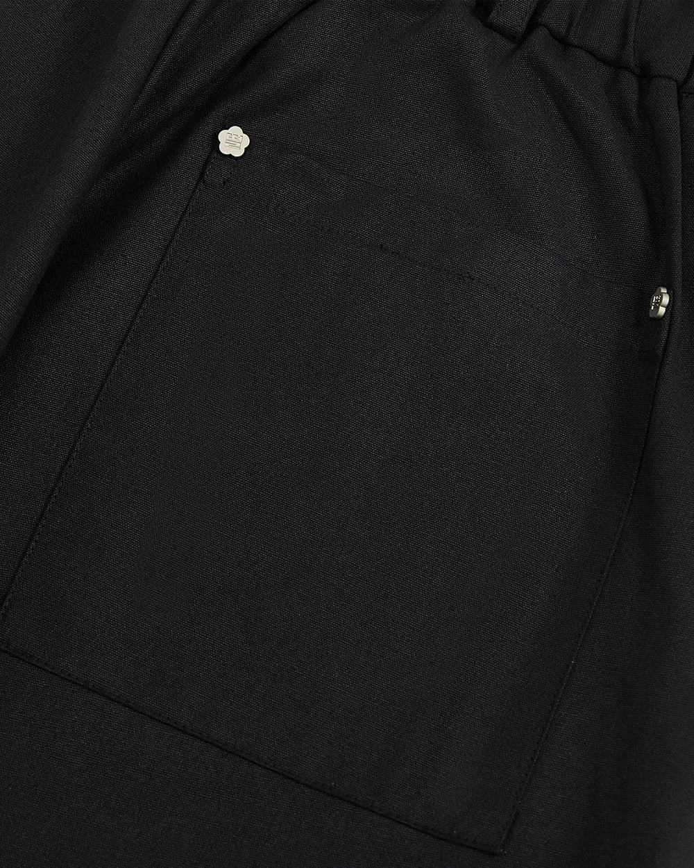 Close-up of a black fabric pocket with two metal rivets—one adorned with a small flower-shaped embellishment—at the top corner, adding a delicate accent to the BLACK HANBOK BALLOON PANTS by Sundae School.