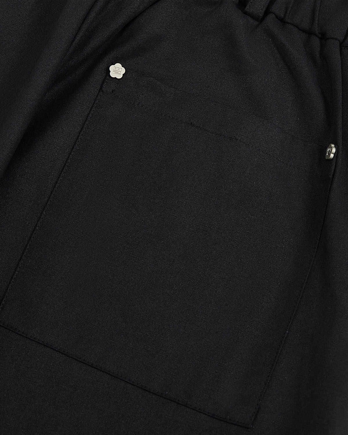 Close-up of a black fabric pocket with two metal rivets—one adorned with a small flower-shaped embellishment—at the top corner, adding a delicate accent to the BLACK HANBOK BALLOON PANTS by Sundae School.