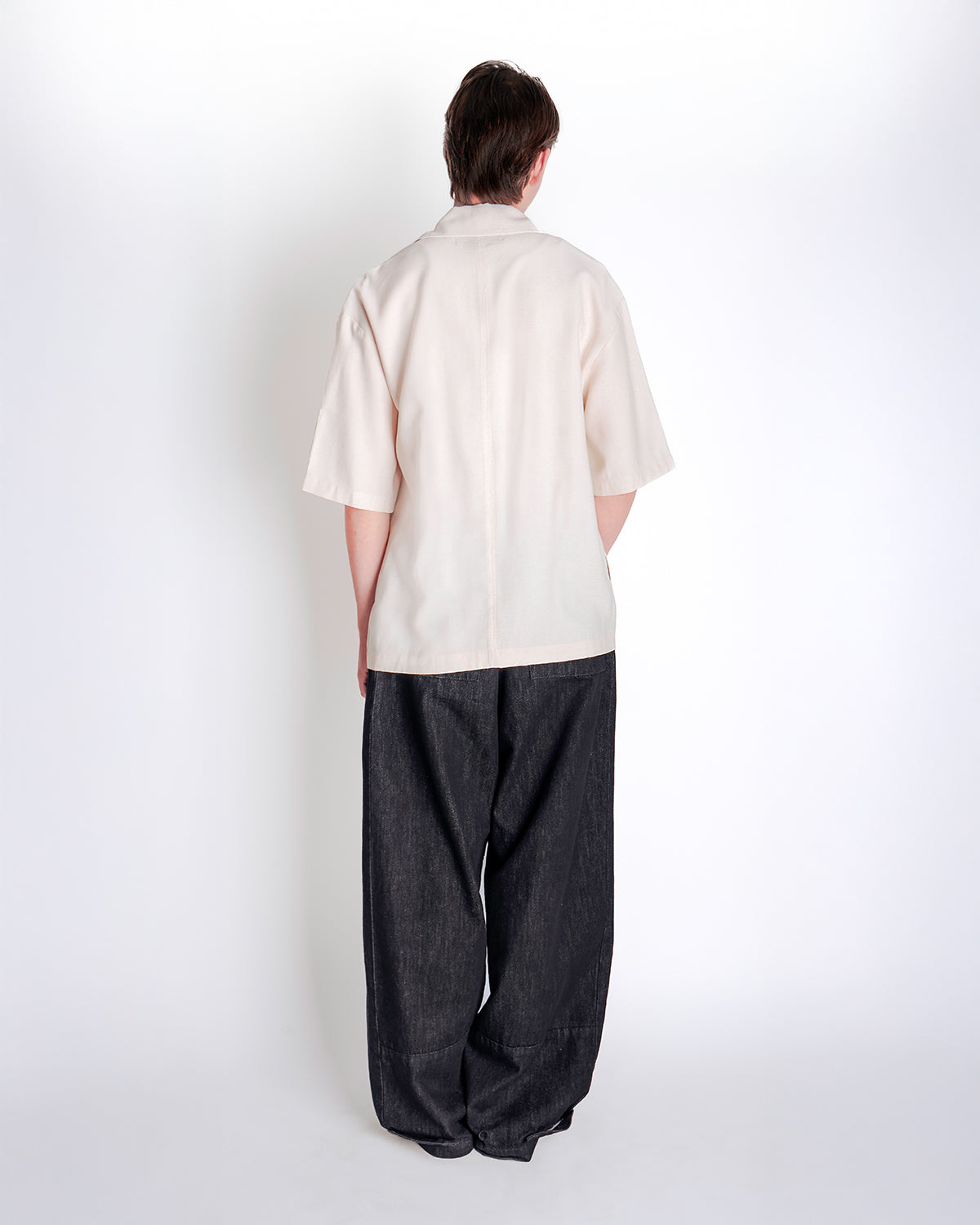 Back view of male model wearing cream hanbok-inspired short sleeve shirt with black wide-leg pants against white studio background, showing relaxed fit and back construction