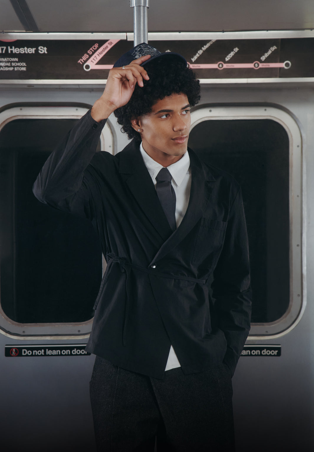 A man wearing a black belted wrap jacket, white dress shirt, and dark tie stands on a subway train near a window.