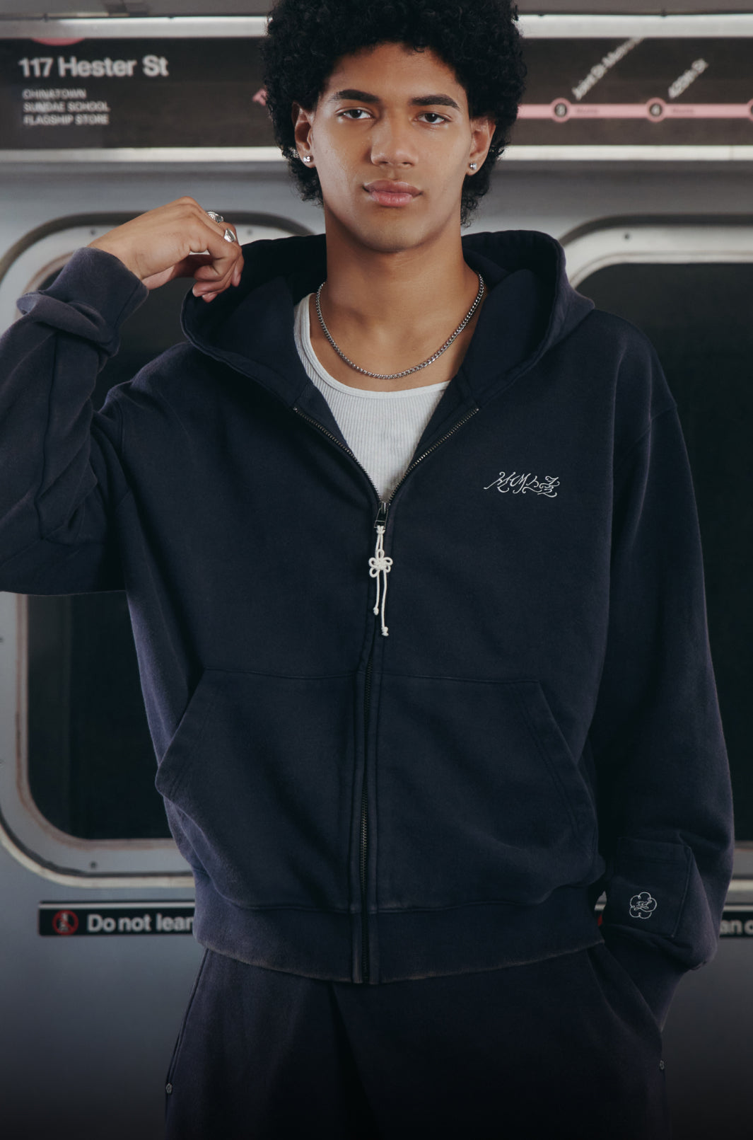 A man wearing a dark navy zip-up hoodie with a small white script logo on the left chest, layered over a white t-shirt and black sweatpants, standing in front of a subway train window.