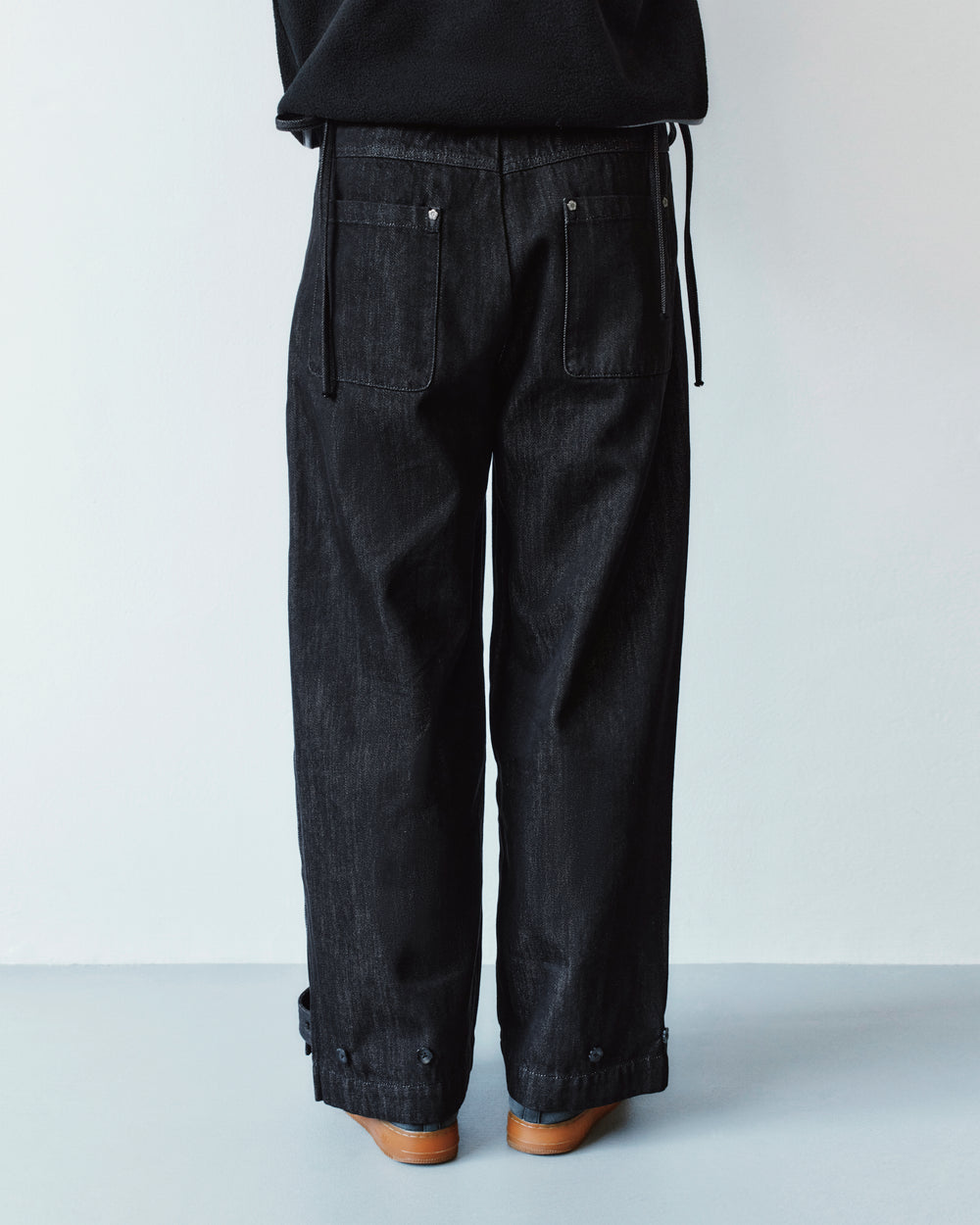 Back view of Sundae School BLACK ACCORDION DENIM PANTS in black denim, featuring rear patch pockets, inverted pleats, and button-tab cuffs at the ankles.