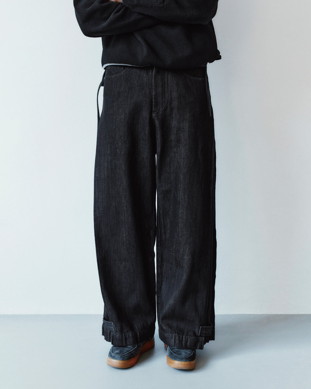 A person wearing loose-fit black BLACK ACCORDION DENIM PANTS with inverted pleats, cuffed hems, and dark loafers.