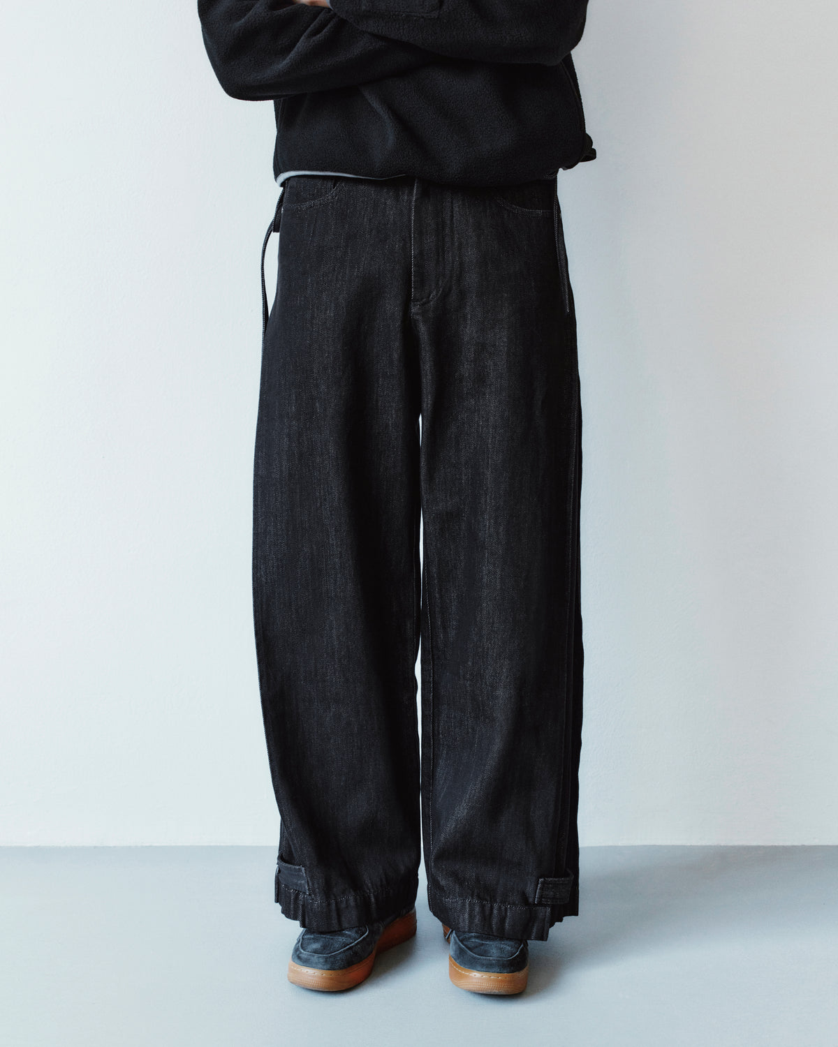 A person wearing loose-fit black BLACK ACCORDION DENIM PANTS with inverted pleats, cuffed hems, and dark loafers.
