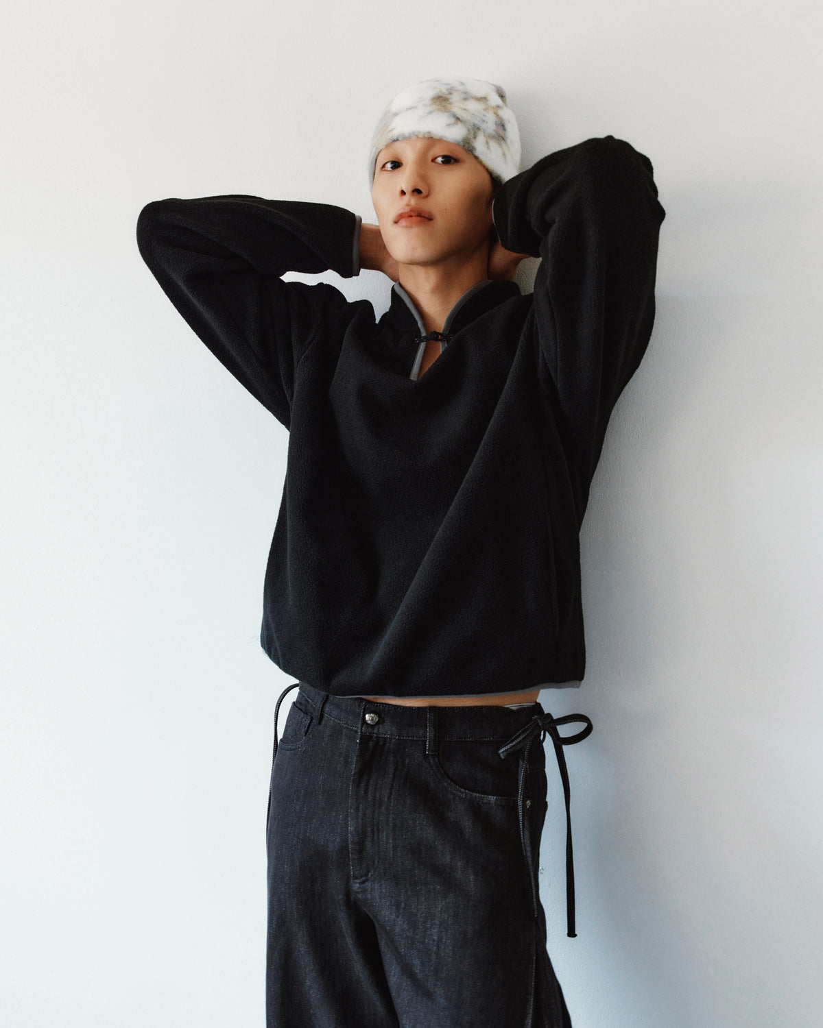 A person wearing a black fleece pullover and BLACK ACCORDION DENIM PANTS with side ties stands against a white wall.