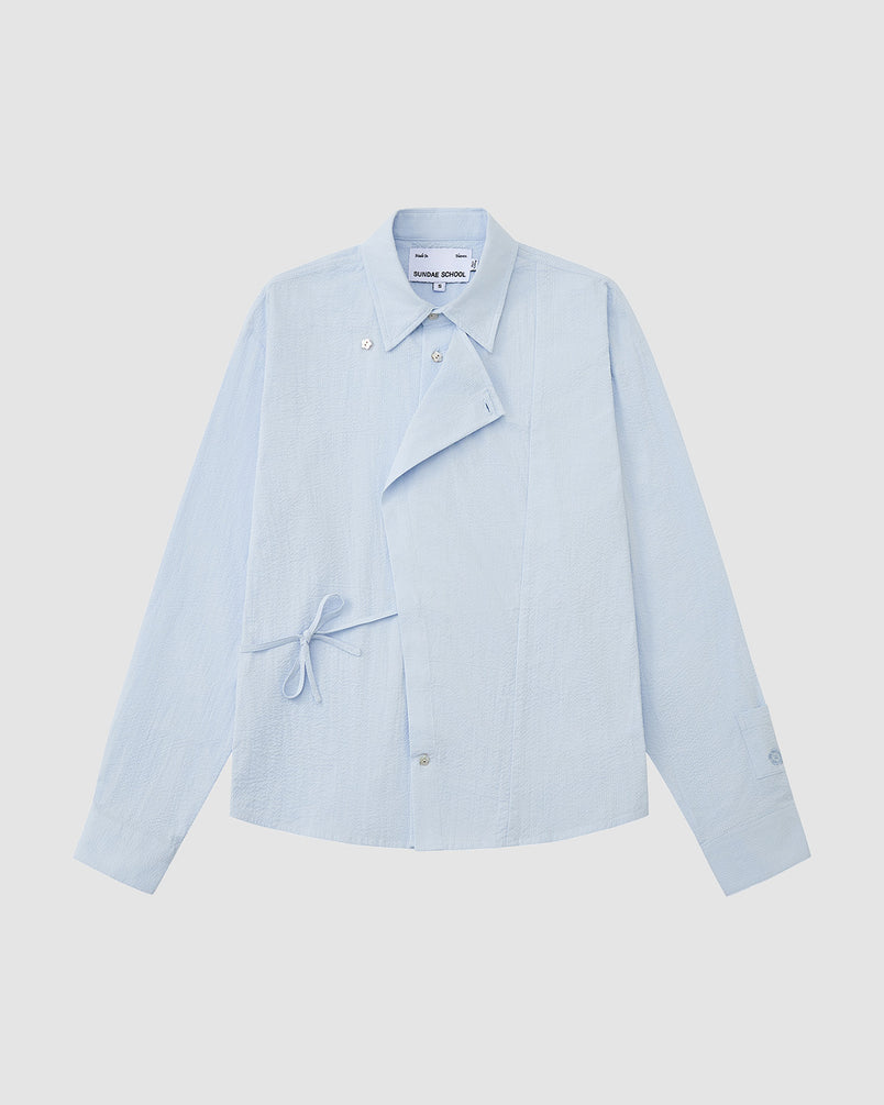 Sundae School SKY BLUE TIE GORUM SHIRT: light blue seersucker cotton, long sleeves, collar, asymmetric buttoned flap with gorum tie, single chest pocket, and buttoned cuffs. Shown on a plain white background.