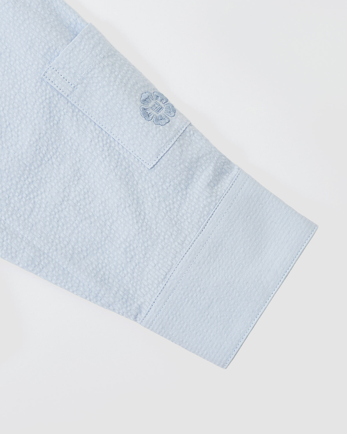 A close-up of the SKY BLUE TIE GORUM SHIRT by Sundae School shows its textured light blue seersucker cotton sleeve cuff, featuring a rectangular patch and an embroidered floral emblem, laid flat on a white background.