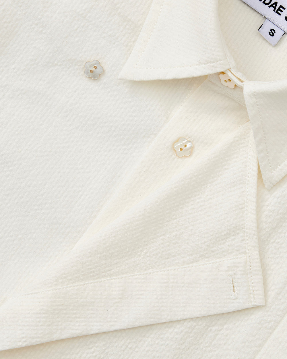 Close-up of the IVORY TIE GORUM SHIRT by Sundae School, showcasing its white seersucker texture, pointed collar, pearlescent buttons, neat stitching, and a partially visible size S tag near the collar.