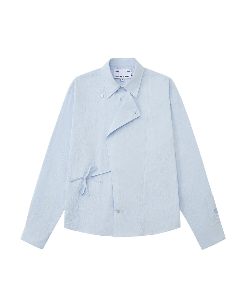 A front flat-lay view of a light blue long-sleeve button-up shirt in a textured seersucker fabric. It features a Korean Hanbok-inspired asymmetrical wrap-front flap, a side tie closure at the waist, a pointed collar, and subtle flower-shaped button accents.