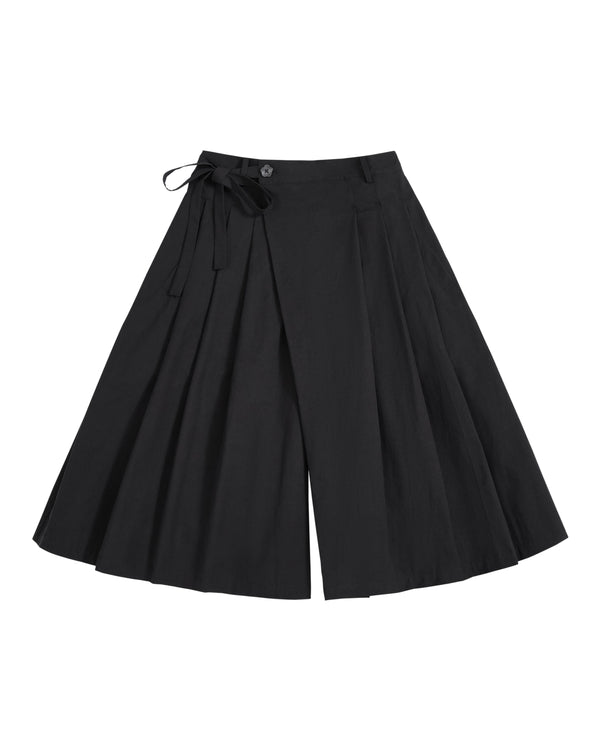 The ASYMMETRIC CHIMA CULOTTE by Sundae School, featuring black wide-leg pleated pants with a buttoned waist and a tied ribbon detail on one side, evokes the flowing elegance of a hanbok chima and is displayed laid flat on a white background.