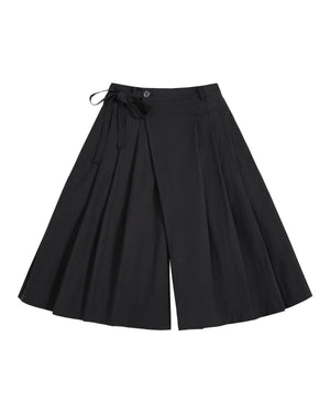 The ASYMMETRIC CHIMA CULOTTE by Sundae School, featuring black wide-leg pleated pants with a buttoned waist and a tied ribbon detail on one side, evokes the flowing elegance of a hanbok chima and is displayed laid flat on a white background.